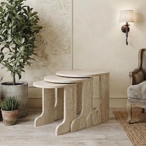 May include: A set of three nesting tables crafted from light beige travertine stone, each featuring a smooth, rounded tabletop and a sturdy, curved base. The tables are arranged in a cascading formation, showcasing the natural porous texture.