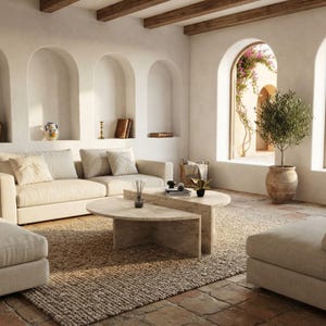 May include: A beige fabric sofa with matching cushions, a two-piece semi-circular stone coffee table, and a potted olive tree in a terracotta vessel. The room features arched wall niches, exposed wooden ceiling beams, and a textured woven rug.