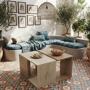Può includere: Un divano angolare in cemento chiaro con cuscini a motivi blu e una coperta, abbinato a due tavolini da caffè cubici in legno chiaro. Lo spazio presenta una lampada a sospensione in vimini, ulivi e agrumi in vaso e stampe botaniche.