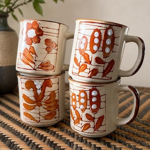 May include: Four ceramic mugs with a cream-coloured base and brown hand-painted floral patterns. Each mug features a grid-like design with orange-brown botanical motifs and small white dot accents. The mugs have sturdy handles and a cylindrical shape.