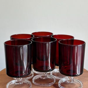 May include: A set of six matching drinking glasses featuring deep red, translucent glass bowls with straight sides. Each glass is supported by a clear, circular glass stem and a wide, flat, clear glass base, creating a uniform silhouette.