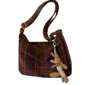 May include: A hobo-style shoulder bag featuring a dark red, green, and yellow plaid pattern. The bag includes an adjustable strap, a gold-toned oval brand plaque, and a plush brown rabbit charm attached to the side with a gold-colored metal clip.