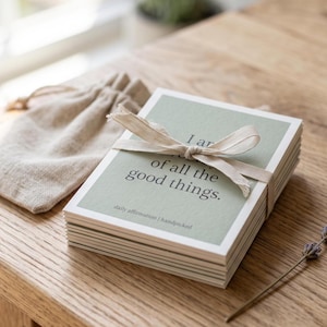 Könnte beinhalten: Ein Stapel rechteckiger Karten mit salbeigrünen Rändern und weißen Mitten, zusammengebunden mit einem beigen Band. Die oberste Karte zeigt den Text "I am worthy of all the good things." und "daily affirmation | handpicked" in schwarzer Schrift, dazu ein Leinenbeutel.