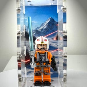 May include: A LEGO minifigure dressed in an orange flight suit with a white helmet featuring a red Rebel Alliance symbol. The figure holds a translucent blue lightsaber and a black blaster, displayed inside a clear acrylic case with a snowy mountain backdrop.