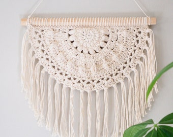 Puede incluir: Un tapiz de pared de crochet blanco con un diseño circular y flecos. El tapiz está sujeto a una varilla de madera.