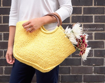 May include: A yellow crocheted tote bag with wooden handles. The bag is filled with white and red flowers. The bag is being held by a person wearing a white shirt and blue jeans.