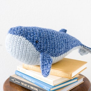 May include: A blue and gray crocheted whale plush toy sitting on a stack of books. The whale has a small black eye and a textured surface.