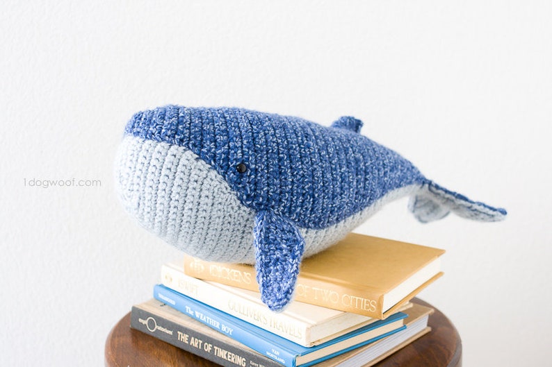 May include: A blue and gray crocheted whale plush toy sitting on a stack of books. The whale has a small black eye and a textured surface.