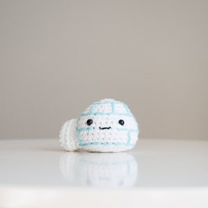May include: A white crocheted snowman with a blue outline and black eyes and mouth. The snowman is sitting on a white surface.