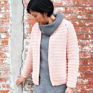 May include: A light pink, open-front crochet cardigan. The handmade sweater has long sleeves and a textured stitch pattern. The model is wearing a gray turtleneck and a gray dress. The background is a brick wall.