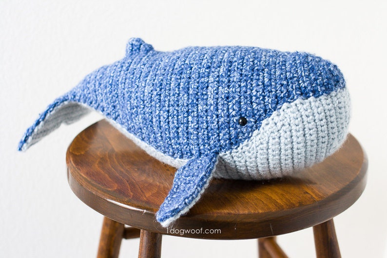 May include: A crocheted blue whale stuffed animal with a light blue belly, sitting on a wooden stool.