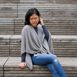 Peut inclure: Une femme portant un châle en tricot gris, un jean bleu avec un genou usé et des bottines marron est assise sur des marches en bois.