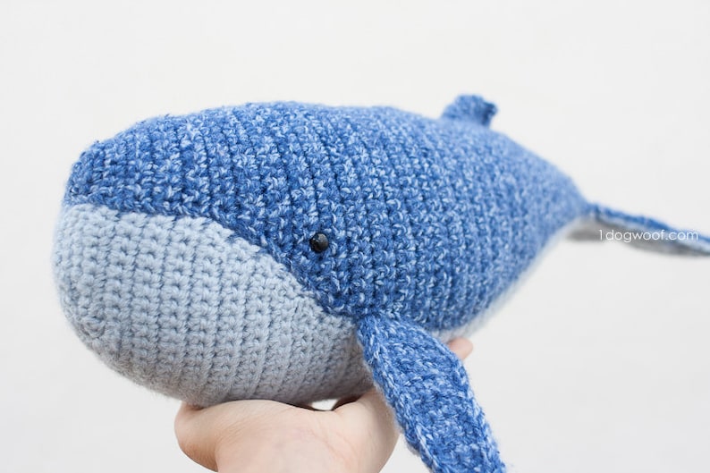 May include: A crocheted blue whale plush toy with a light blue belly. The whale has a black eye and is held in a person's hand.