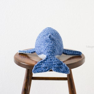 May include: A blue and white crocheted whale plush toy sitting on a wooden stool. The whale is facing away from the camera, with its tail facing the viewer.