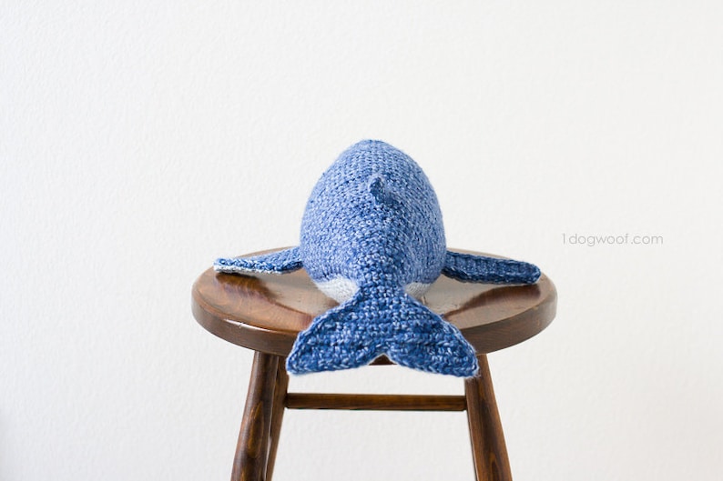 May include: A blue and white crocheted whale plush toy sitting on a wooden stool. The whale is facing away from the camera, with its tail facing the viewer.