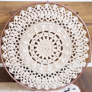 May include: A cream-colored, circular crochet doily, suspended within a copper-colored metal ring. The intricate, handmade design features a central floral pattern and a scalloped edge. This decorative piece is suitable for wall decor.