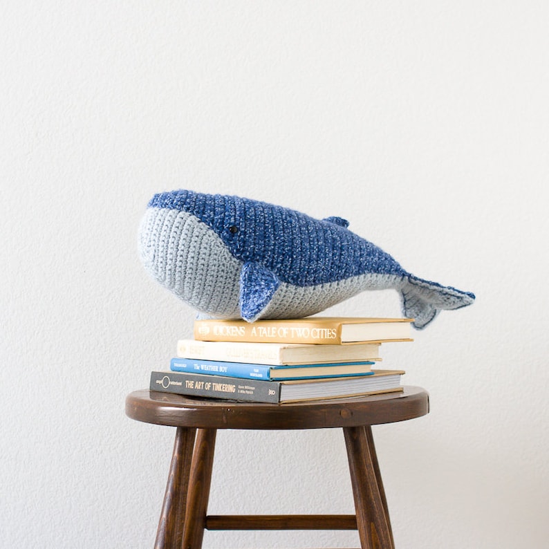 May include: A blue and gray crocheted whale toy sitting on top of a stack of books on a wooden stool. The books are titled "A Tale of Two Cities", "The Weather Boy", and "The Art of Tinkering".