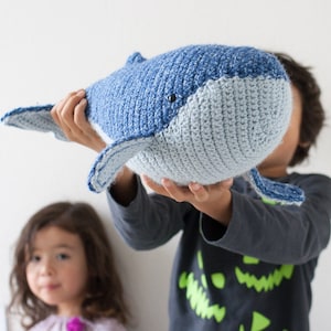 May include: A crocheted blue and gray whale plush toy being held by a child. The whale has a stitched-on eye and a small mouth.