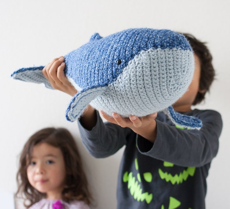 May include: A crocheted blue and gray whale plush toy being held by a child. The whale has a stitched-on eye and a small mouth.