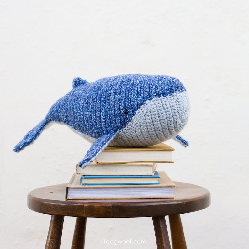 May include: A blue and gray crocheted whale plush toy sitting on top of a stack of books on a wooden stool.