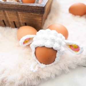 May include: A white crocheted lamb costume for an egg, with pink and yellow ears. The costume is sitting on a white fluffy surface with other eggs in the background.