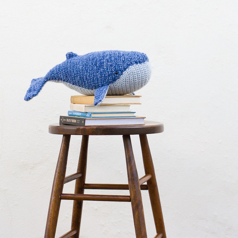 May include: A blue crocheted whale plush toy sitting on top of a stack of books on a wooden stool.