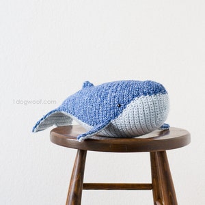 May include: A crocheted blue and gray whale plush toy sitting on a wooden stool. The whale has a simple face with black eyes.