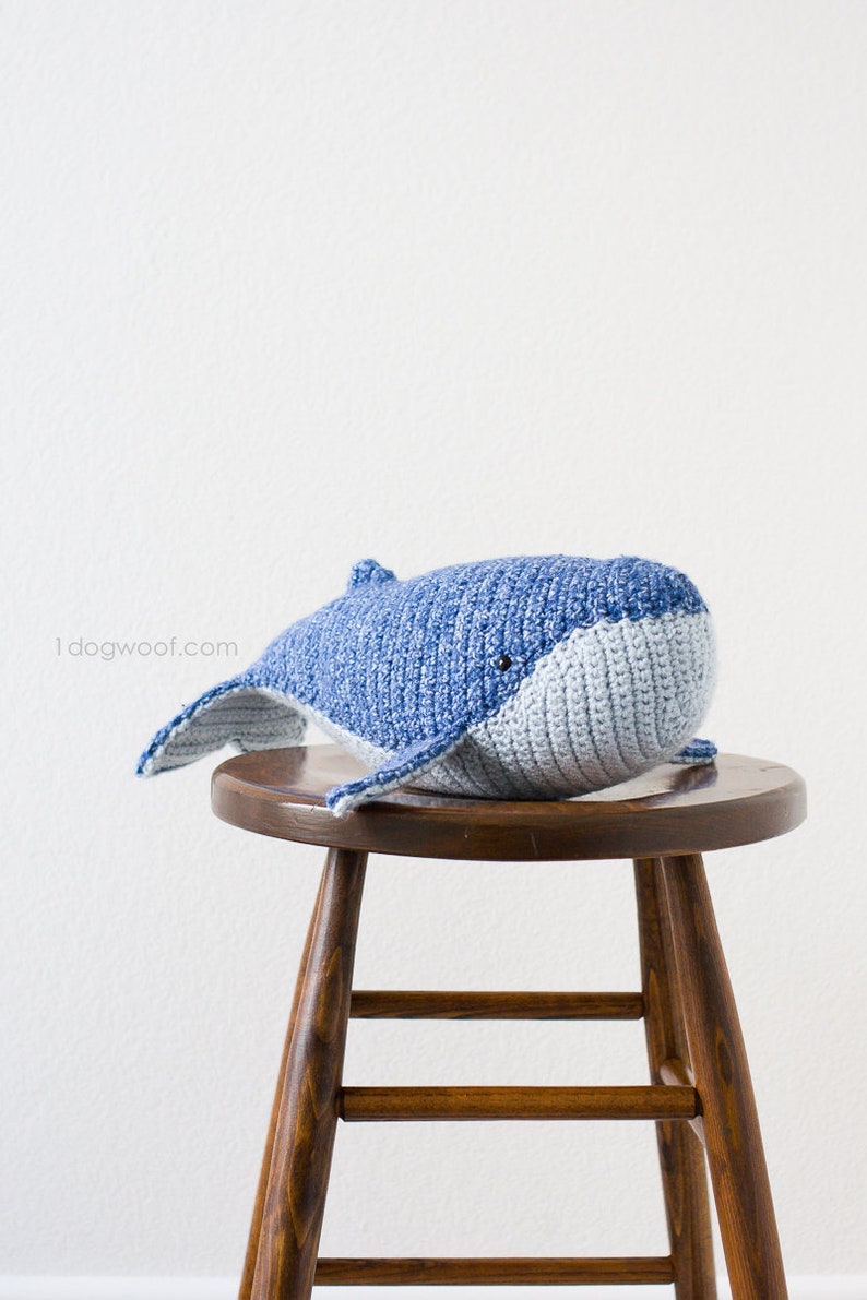 May include: A crocheted blue and gray whale plush toy sitting on a wooden stool. The whale has a simple face with black eyes.