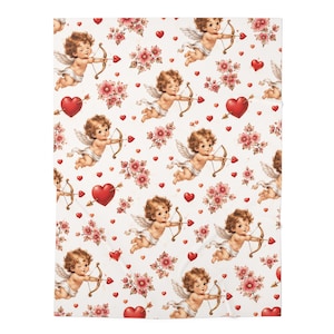May include: A white fabric blanket featuring a repeating pattern of cherubic figures with wings, each holding a bow and arrow. The design includes scattered red hearts and clusters of small pink flowers, creating a dense, decorative motif across the surface.