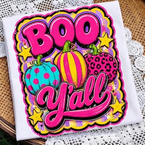 Peut inclure: Un t-shirt blanc avec un motif graphique vibrant portant le texte BOO Y'ALL en rose vif. Le design comprend trois citrouilles : une sarcelle à pois roses, une rayée jaune et rose, et une rose avec un motif imprimé léopard noir.