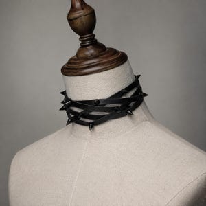 Könnte beinhalten: Ein schwarzes Multi-Wrap-Halsband, bestehend aus dünnen, lederartigen Riemen, die mehrfach um den Hals gewickelt sind. Das Design umfasst mehrere scharfe, spitze schwarze Metallnieten, die gleichmäßig entlang der Bänder verteilt sind.
