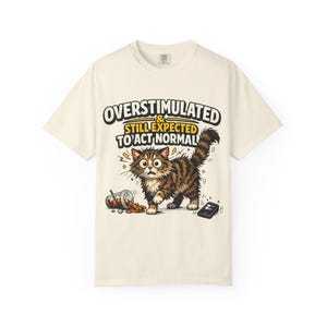 May include: A cream-colored t-shirt featuring a graphic of a wide-eyed, fluffy brown tabby cat. The text reads OVERSTIMULATED & STILL EXPECTED TO ACT NORMAL. A spilled plastic cup with a straw and a black smartphone lie on the ground near the cat.