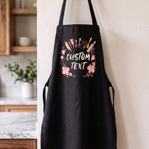 May include: A black apron featuring a central graphic with the words CUSTOM TEXT surrounded by a wreath of pink flowers and various kitchen utensils including a rolling pin, whisk, and spatula arranged in a circular pattern on the chest area.