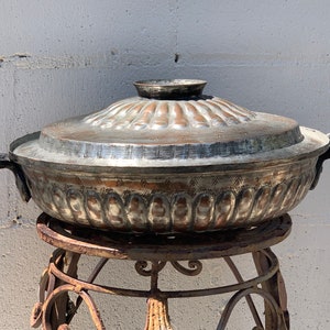 May include: A large, round, silver-colored copper pot with a lid. The pot has a decorative design around the rim and is sitting on a metal stand.