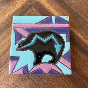 May include: A square ceramic tile with a black bear silhouette on a blue, purple, and pink geometric background.