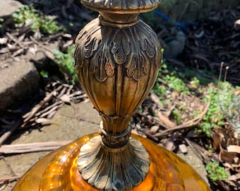 May include: Close-up of a vintage amber glass lamp with a brass-colored metal base. The lamp features ornate detailing on the metal components and a large, rounded glass shade. The lamp is designed to be a decorative lighting fixture.