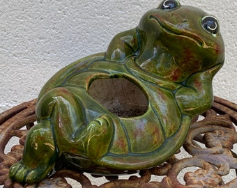 Puede incluir: Una maceta de cerámica con forma de rana verde, apoyada sobre una mesa de metal decorativa. La rana tiene una cara sonriente y está en una pose relajada. La maceta tiene un área ahuecada para plantas. La mesa tiene un diseño ornamentado.