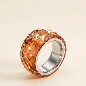 Puede incluir: Un anillo de banda ancha con una capa exterior de resina facetada de color ámbar con inclusiones brillantes. El interior del anillo está forrado con una banda de metal liso y pulido en tono plateado, creando un contraste entre el exterior texturizado y la superficie interior metálica.