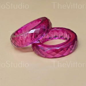 May include: Two vibrant magenta rings featuring a faceted, geometric surface texture. The rings are translucent and catch the light, displaying a deep pink hue throughout their circular bands. The text TheVittorioStudio is visible in the background.