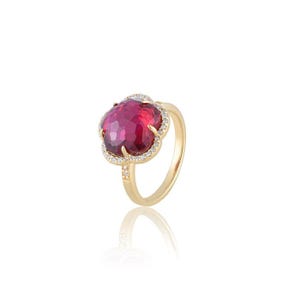 Könnte beinhalten: Ein Goldring mit einem großen, facettierten, runden, dunkelrosa Edelstein in einer blumenförmigen Fassung. Die Fassung ist an den Rändern mit kleinen, klaren, funkelnden Steinen besetzt, und der Ring besteht aus poliertem, glattem Gold.