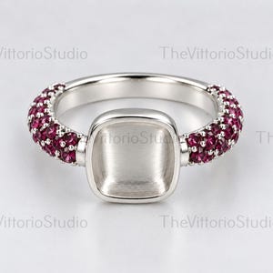 Peut inclure: Une bague en métal argenté avec un chaton carré vide au centre. L'anneau est orné de multiples petites pierres circulaires rose foncé serties en pavé. Le texte VittorioStudio est visible sur l'arrière-plan.