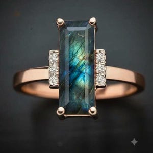 Könnte beinhalten: Ein Ring aus Roségold mit einem rechteckigen, vertikal ausgerichteten Labradorit-Edelstein, der blau und grün schimmert. Der Mittelstein wird von zwei kleinen vertikalen Leisten flankiert, die jeweils mit drei kleinen, klaren Steinen besetzt sind.