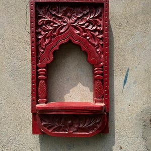 May include: A rectangular, deep red wooden wall shelf featuring an arched opening. The frame is decorated with intricate floral carvings and vertical pillar-like details on the sides. A small, semi-circular shelf base is attached at the bottom.