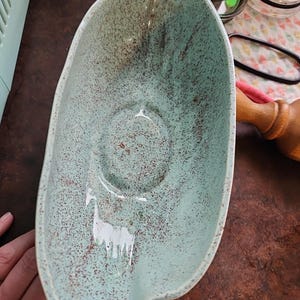 Könnte beinhalten: Eine ovale Keramikschale mit hellminzgrüner Glasur und braun gesprenkeltem Finish. Die Schale hat ein glattes, geschwungenes Inneres mit einer zentralen kreisförmigen Vertiefung und einem leicht unregelmäßigen, organischen Randdesign.
