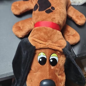 Puede incluir: Un juguete de peluche con forma de perro, con pelaje marrón, orejas largas negras y una nariz negra. El juguete tiene ojos grandes con detalles amarillos y un collar rojo brillante alrededor del cuello. Se ven manchas marrón oscuro en la espalda del peluche.