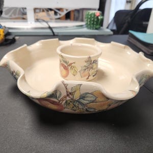 May include: A cream-colored ceramic serving dish with a ruffled edge and a central integrated bowl. The exterior and interior feature a colorful botanical print of various fruits, including pears and apples, with green leaves and brown stems.