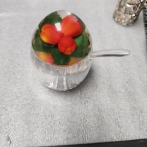 May include: A clear, rounded glass container with a lid featuring an embedded design of bright orange berries and green leaves. The container includes a small, matching clear glass spoon extending from the side, all crafted from smooth, transparent material.