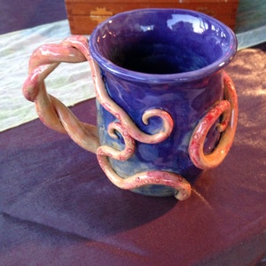 May include: A handmade ceramic mug with a deep blue glaze. The handle is a twisted design, and the mug is adorned with sculpted tentacles in shades of pink and orange. The mug sits on a purple surface.
