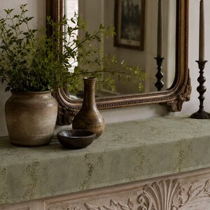 May include: A large, textured, light green fabric runner with a subtle floral pattern draped over a mantelpiece. It features a wide, round, weathered brown ceramic vase filled with green foliage, a tall brown vase, and a small dark brown bowl.