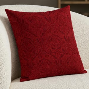May include: A square throw pillow in a deep red color featuring a tonal damask pattern. The fabric displays a repeating intricate botanical motif with swirling leaves and floral shapes woven into the dark red textile surface.